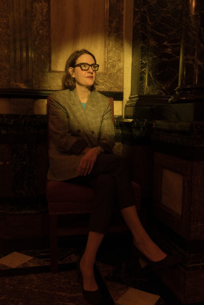 An image of a person sitting down with glasses and a grey blazer, with their hands and legs crossed.