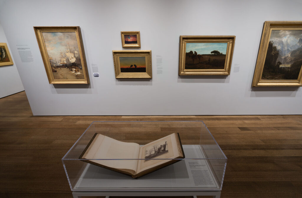 An art gallery featuring framed objects on a white wall and a glass stand with a book in it.