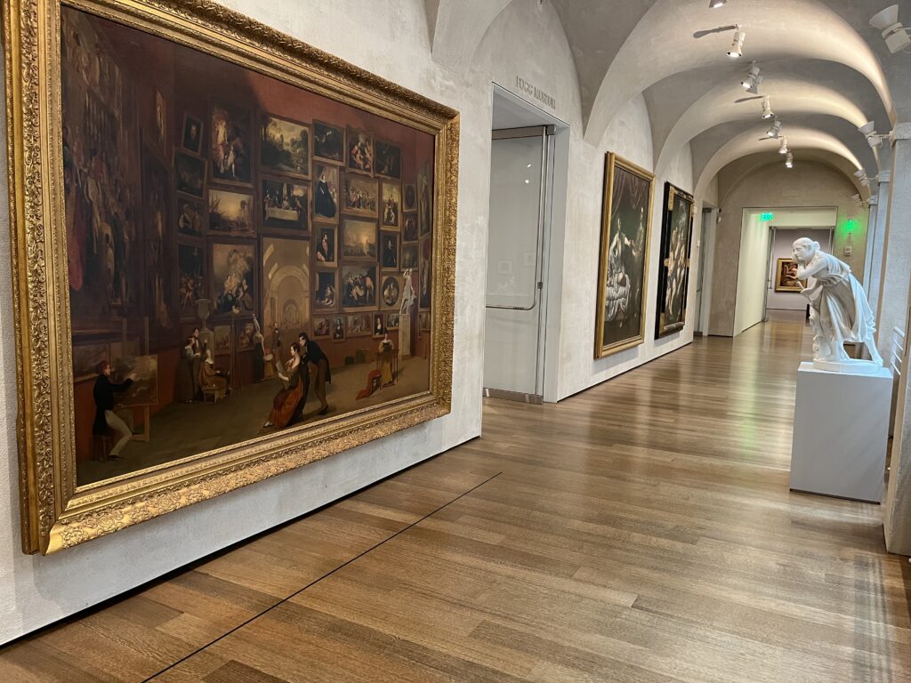 An art gallery hallway with framed objects on one side and a statue-like figure on the other side.