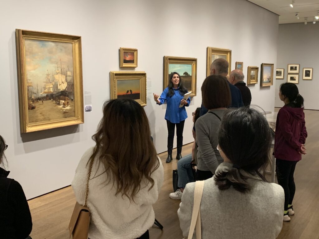 A group of people in an art gallery with a person in a blue shirt leading a discussion.