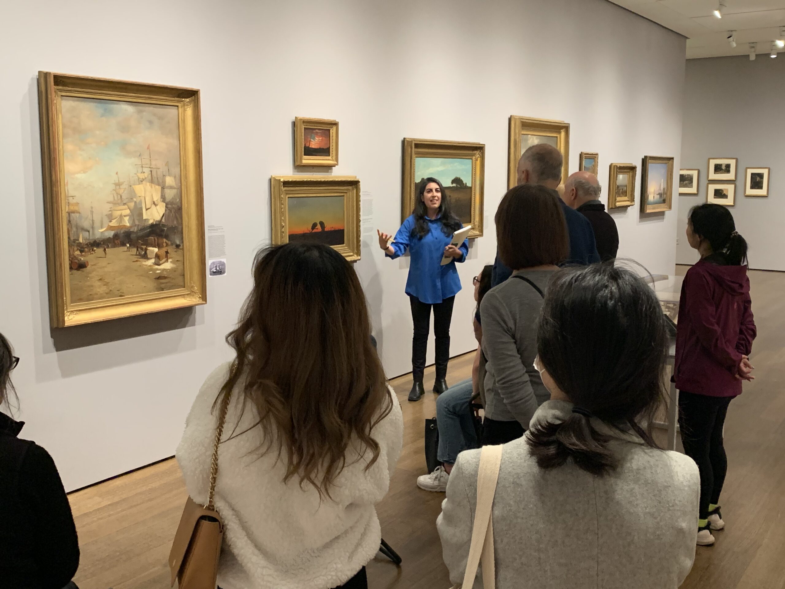 A group of people in an art gallery with a person in a blue shirt leading a discussion.