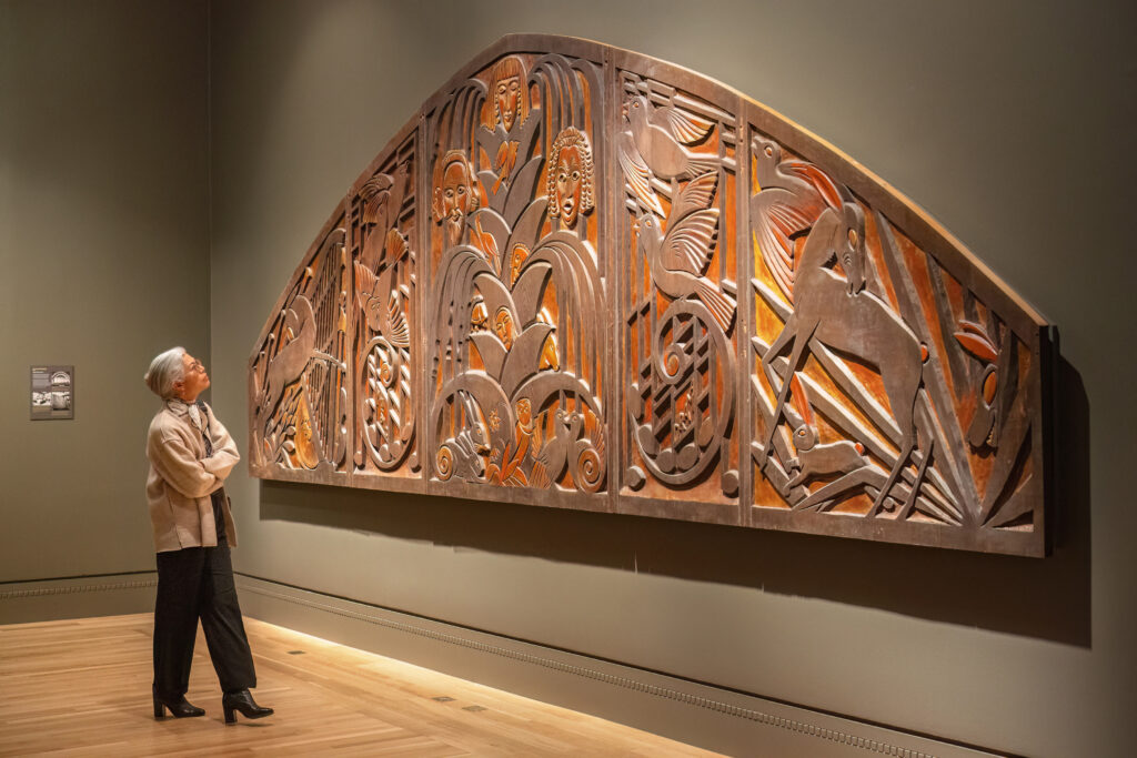 Wood carving installed on a wall. A person looks up at it.