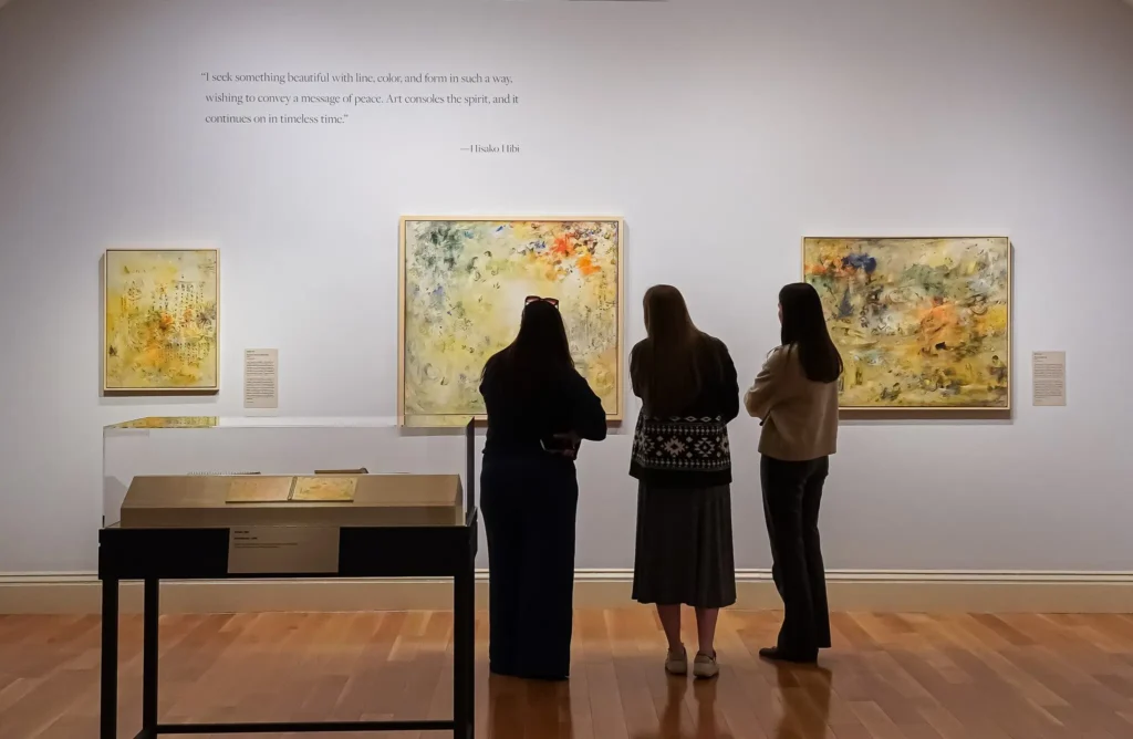 Three people in a gallery looking at three works of art hanging on a wall.