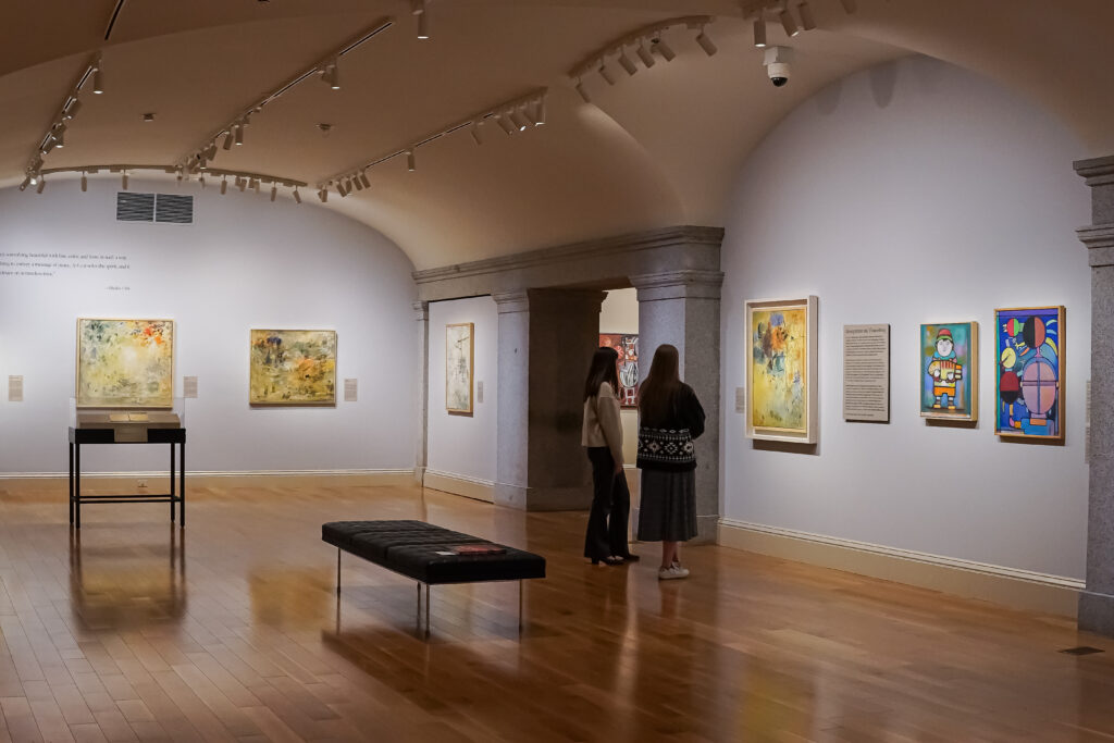 Two people in a gallery space looking a one of several painting hanging on two walls.