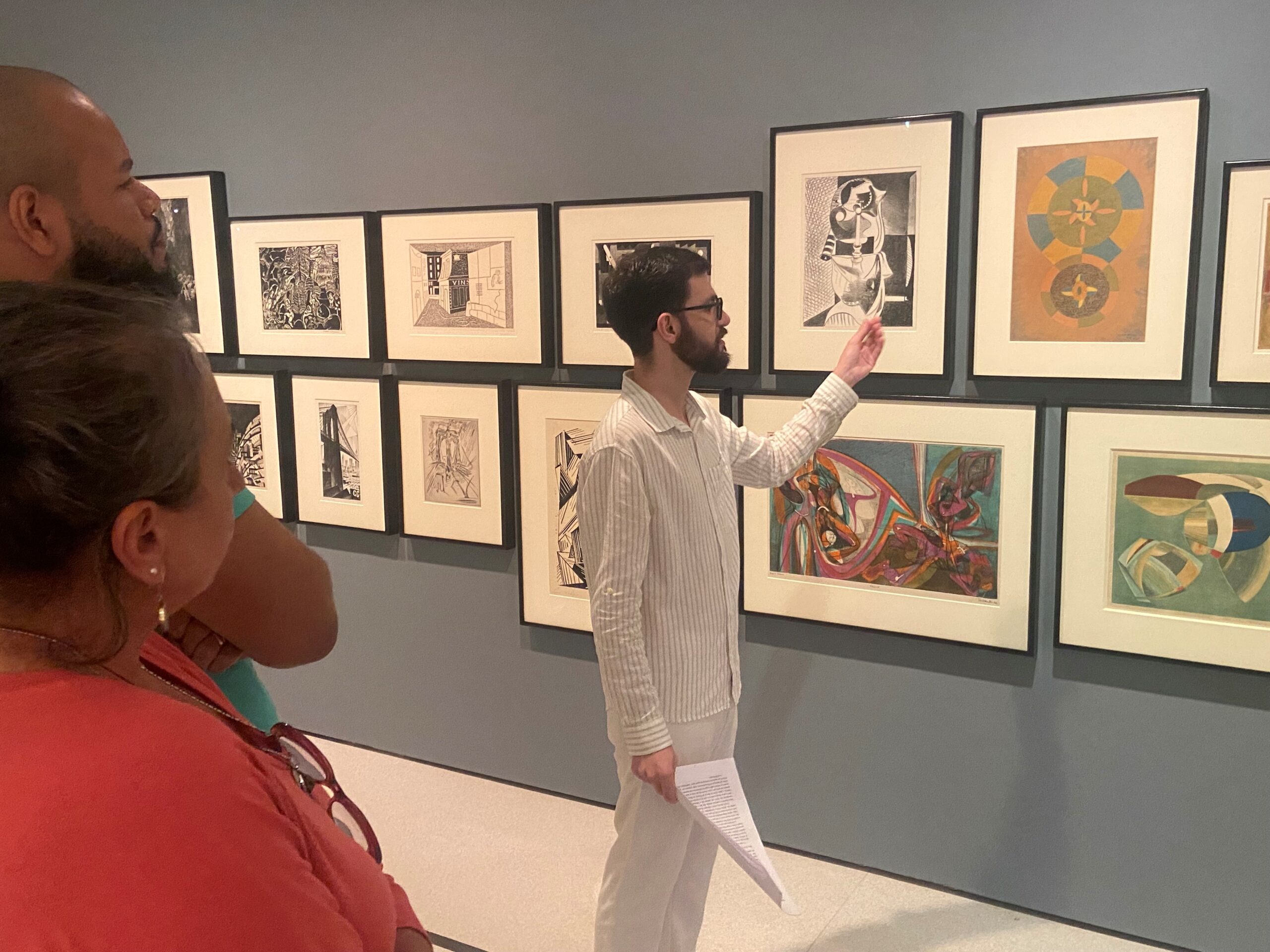 A person standing in an art gallery in front of a wall of many different artworks, pointing specifically at one.