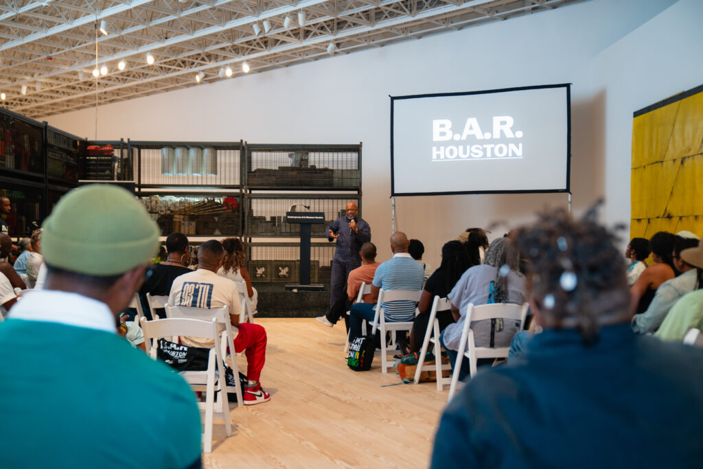 People seated in rows of chairs facing forward, toward Theaster Gates, who is addressing the crowd.