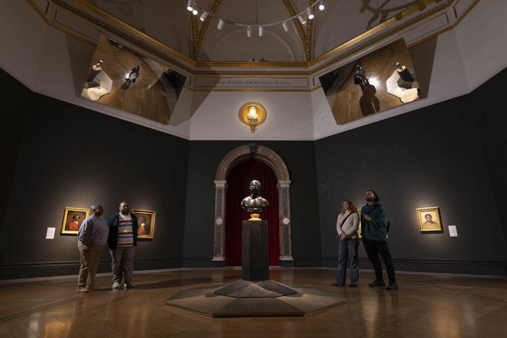Two pairs pf people looking at a sculptural bust on a pedestal in the center of a room.
