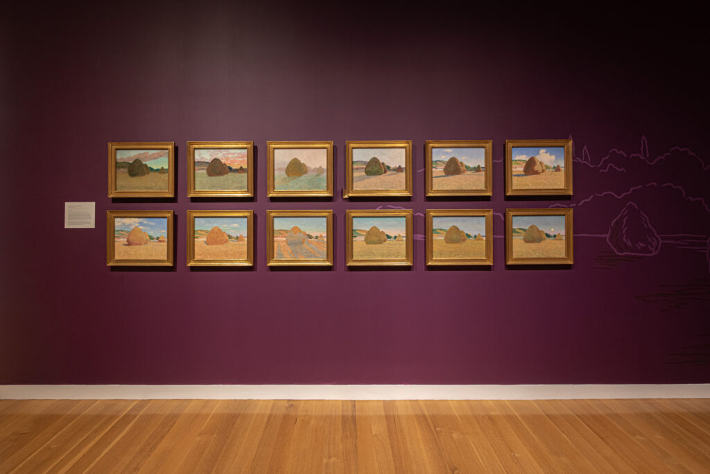 Twelve small paintings, hanging in two rows of six, on a maroon wall.