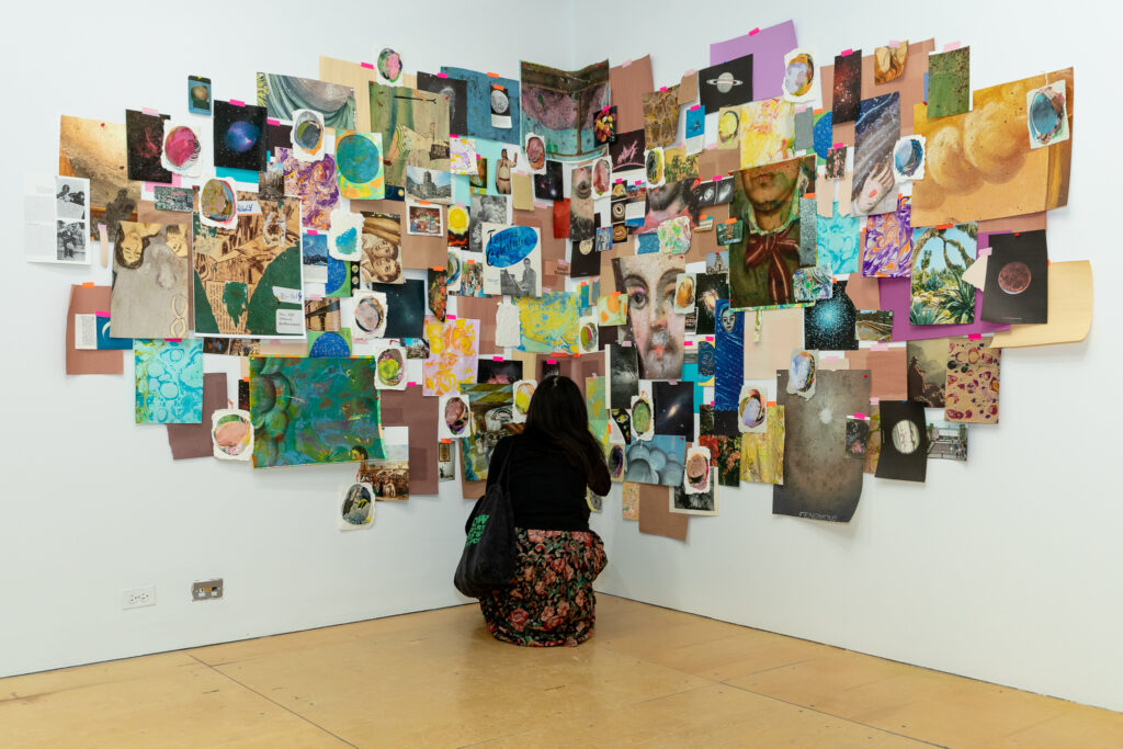 Mixed-media collage installed at the corner of two walls, with a person crouched down, viewing the work.