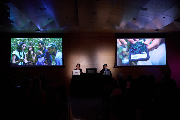 Dark conference room with two people presenting to the audience. Two screens are on either side of the presenters.