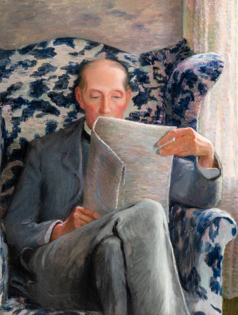 A man dressed in a gray suit sits in a patterned blue-and-white chair by a window and reads a newspaper.