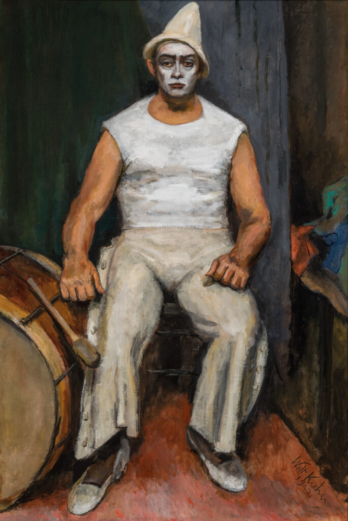 A man dressed as a clown sits in the backstage of a theater next to a large drum.