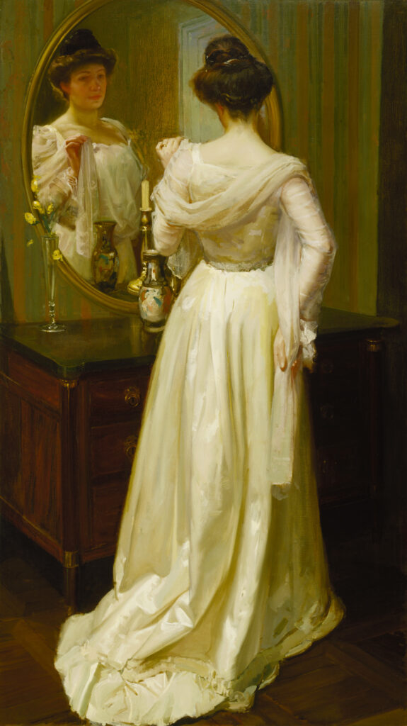 A woman in a white dress wearing a white shawl looks at herself in a mirror.