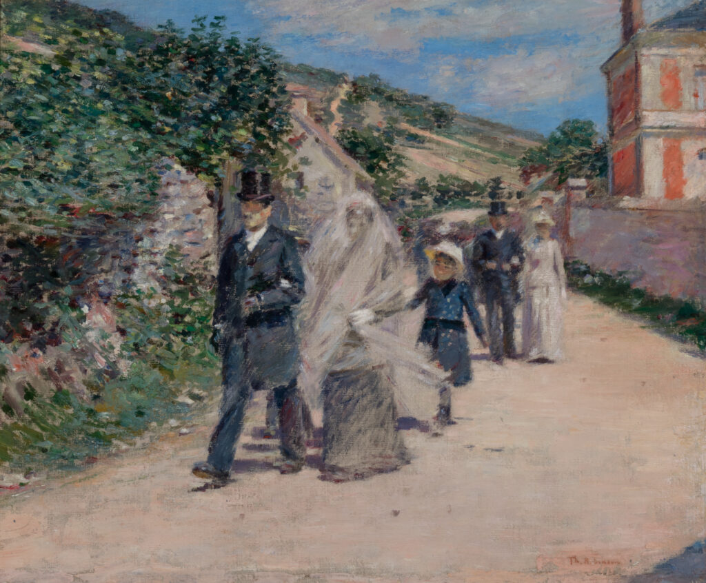 A small group of people walk through a village on a sunny day. The two people in front are dressed in wedding outfits.