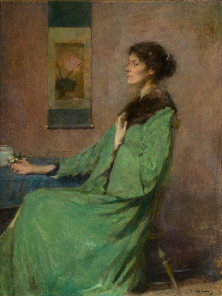 A woman wearing a green dress with a brown fur collar sits in a chair next to a table, resting her right arm on the table.