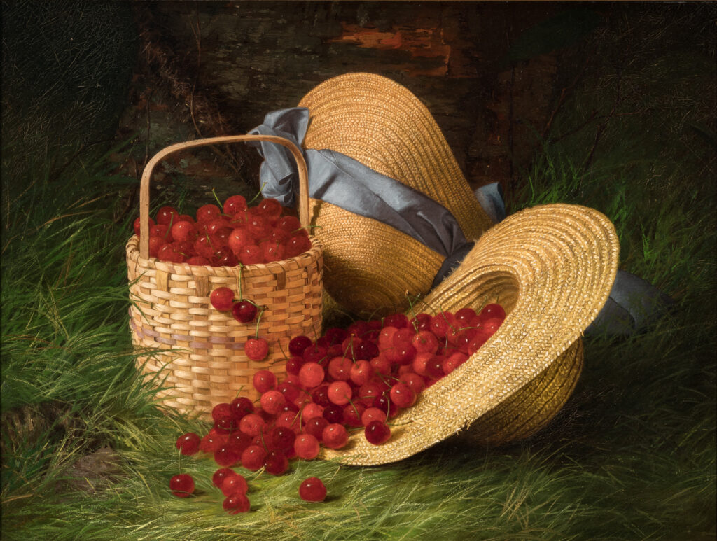 In a patch of tall green grass against a tree trunk, a wicker basket and a straw hat are full of red cherries. A second straw hat with a blue bow rests behind the basket.