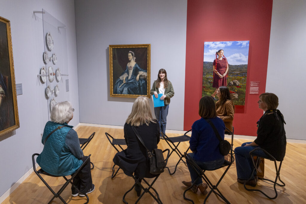 A group of people look at paintings in a gallery. Four people are seated and one person stands.