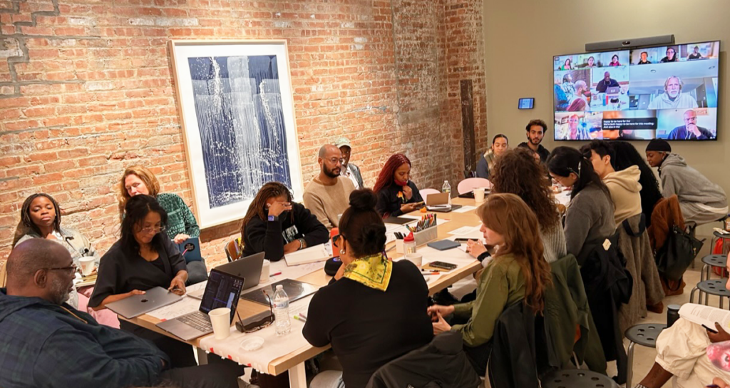 A group of people is seated around a long table with a screen at showing many people on a video call.