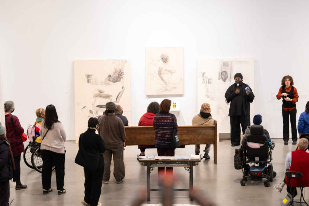 A group of people in an exhibition space listening to one person talking into a microphone. The person talking is standing in front of three works hung on the wall behind.