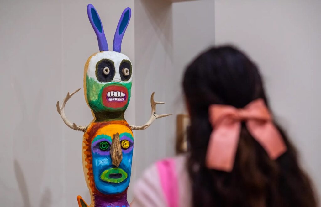 Image of the back of a person's head with a pink bow looking at a painted object with two multi-colored faces.