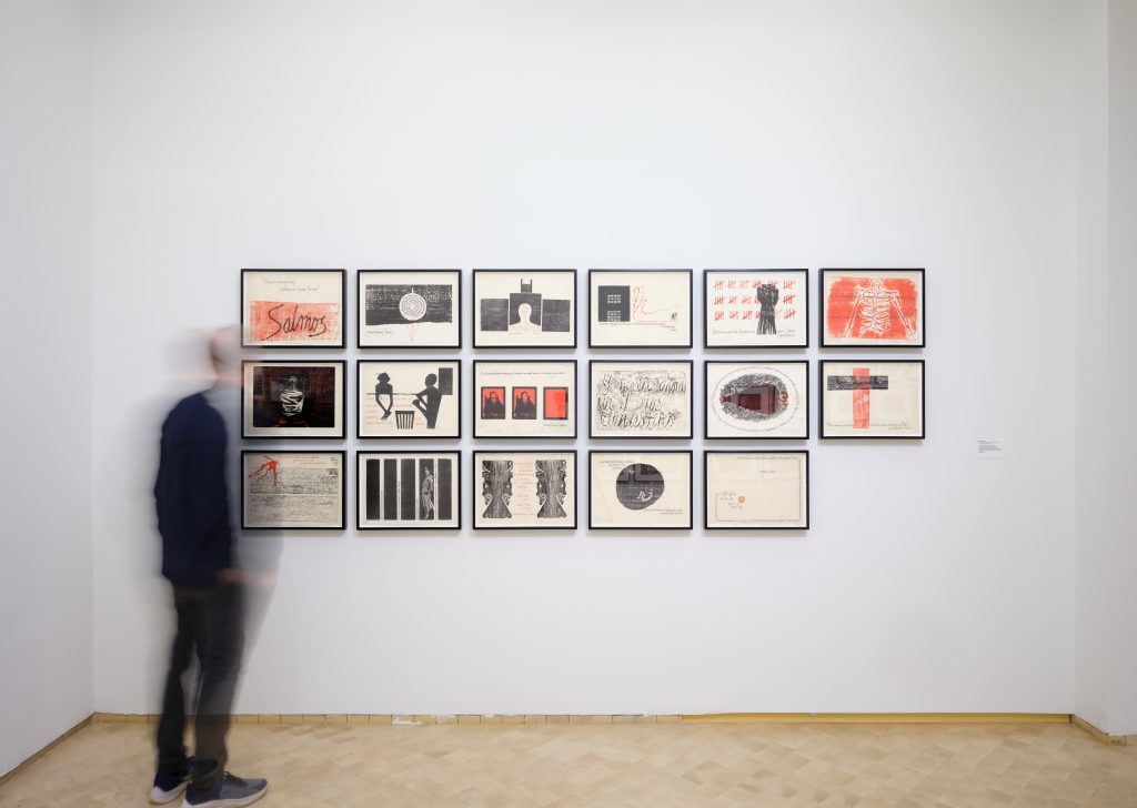 Person looking at three rows of framed works on a wall.