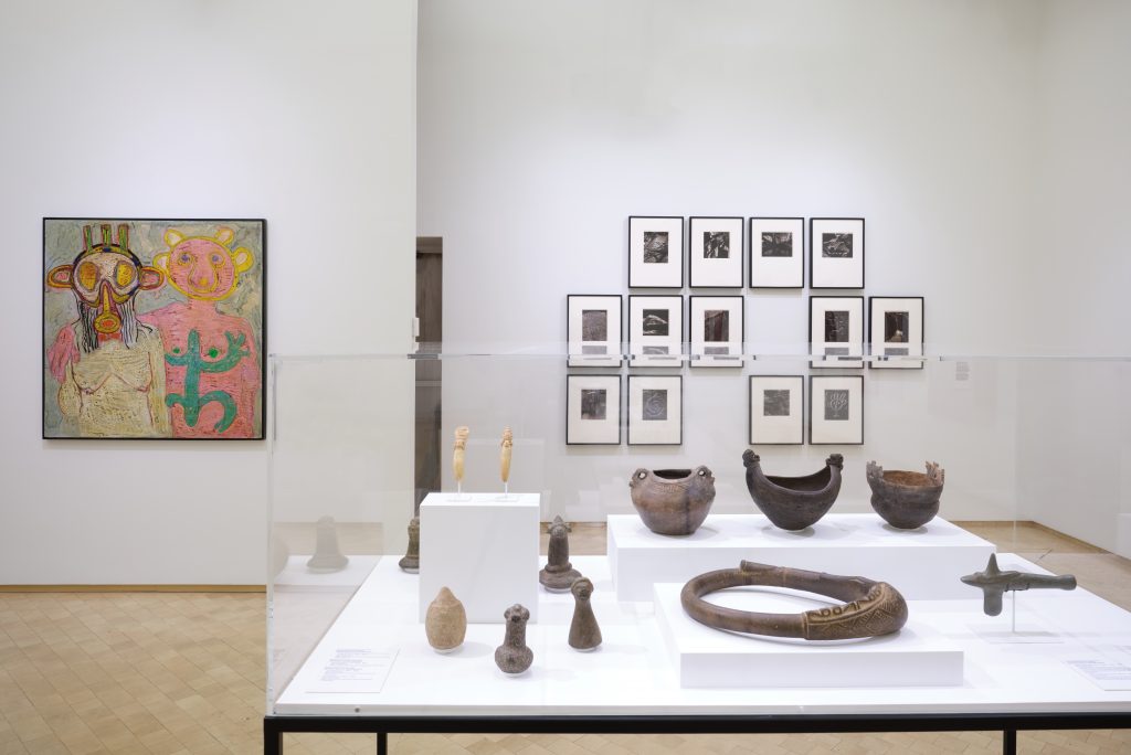 Gallery installation with framed works of art hanging on a wall in the background and a vitrine with objects in the foreground.
