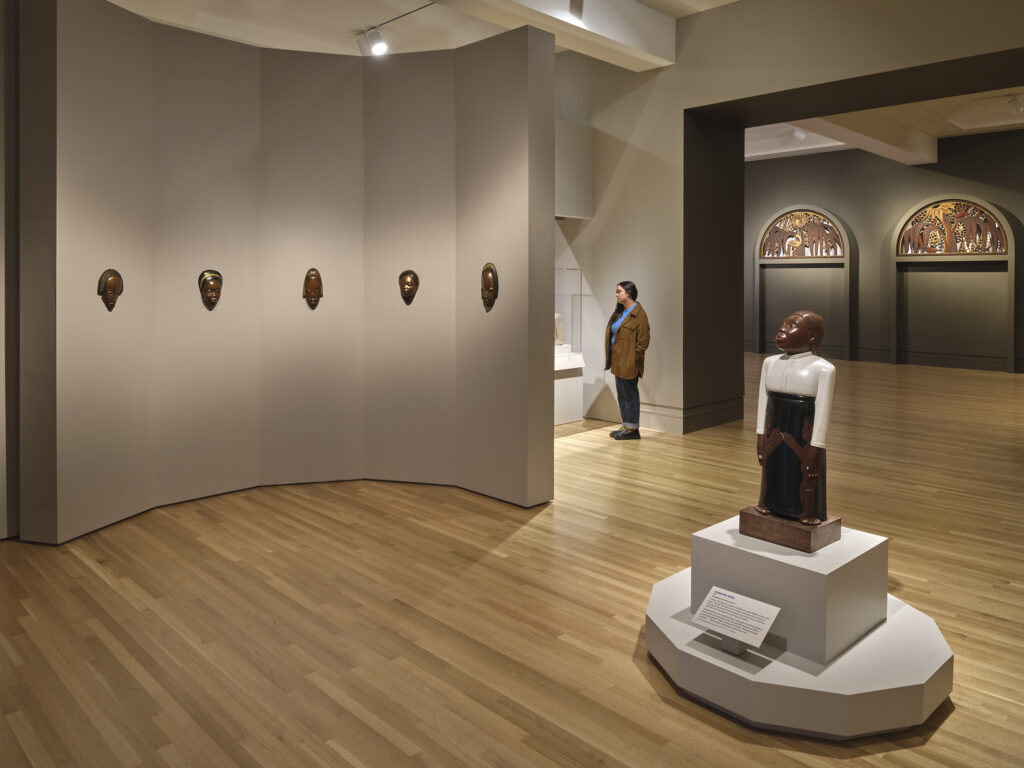 A sculpture in the foreground of a gallery space and a woman standing in the background looking at works of art out of view.