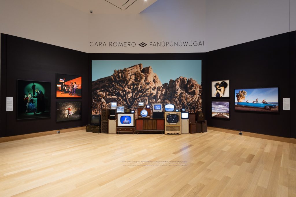 A black gallery wall with works hanging on it and in front of the wall are several old televisions with different images on the screens.