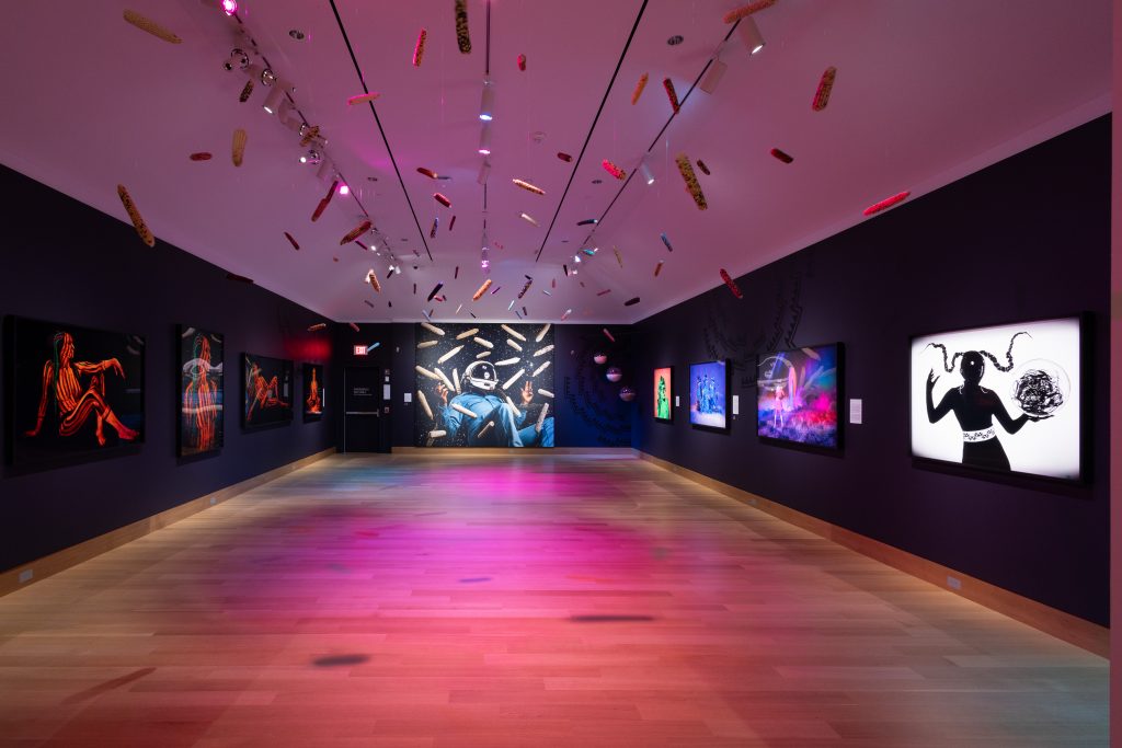 Black gallery walls with works handing an small sculptures suspended from the ceiling bathed in pink light.