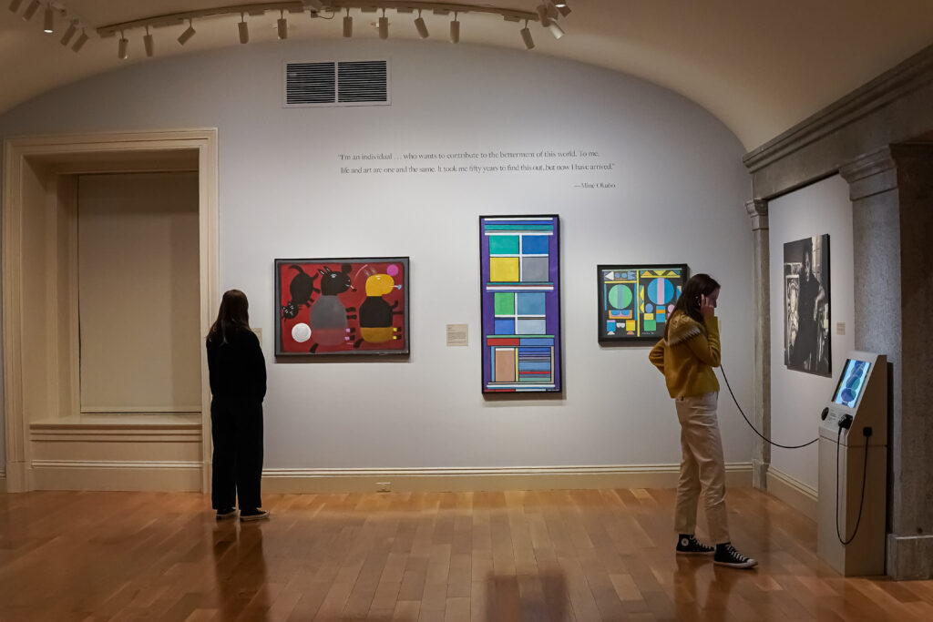 Three framed works hanging on a wall with people standing in front viewing them.