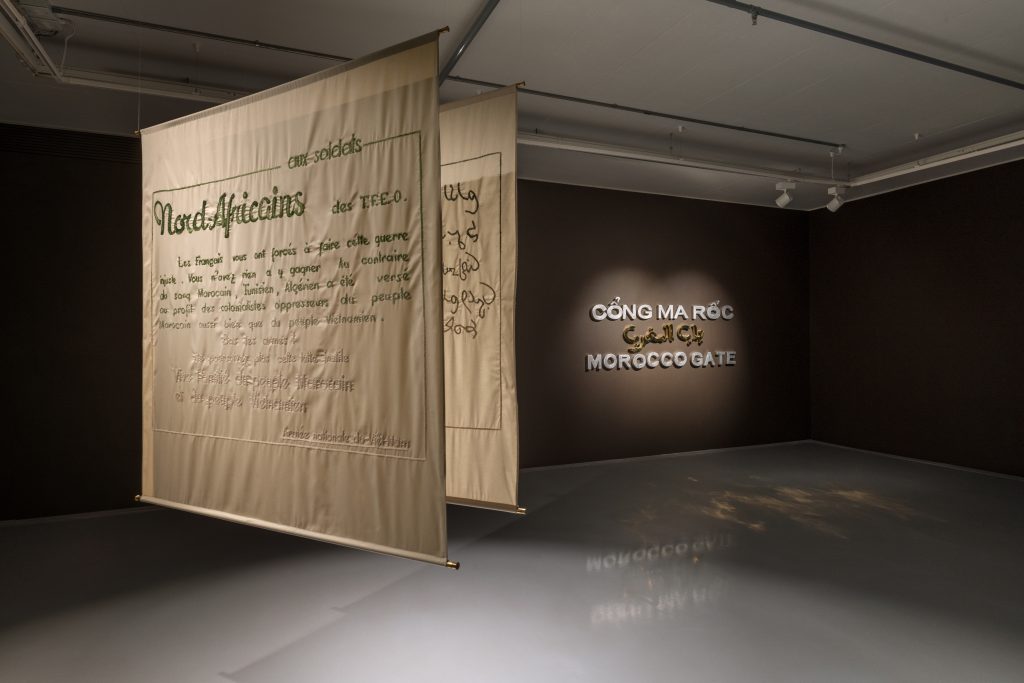 Two textiles suspended from the ceiling with a dark brown title wall behind.