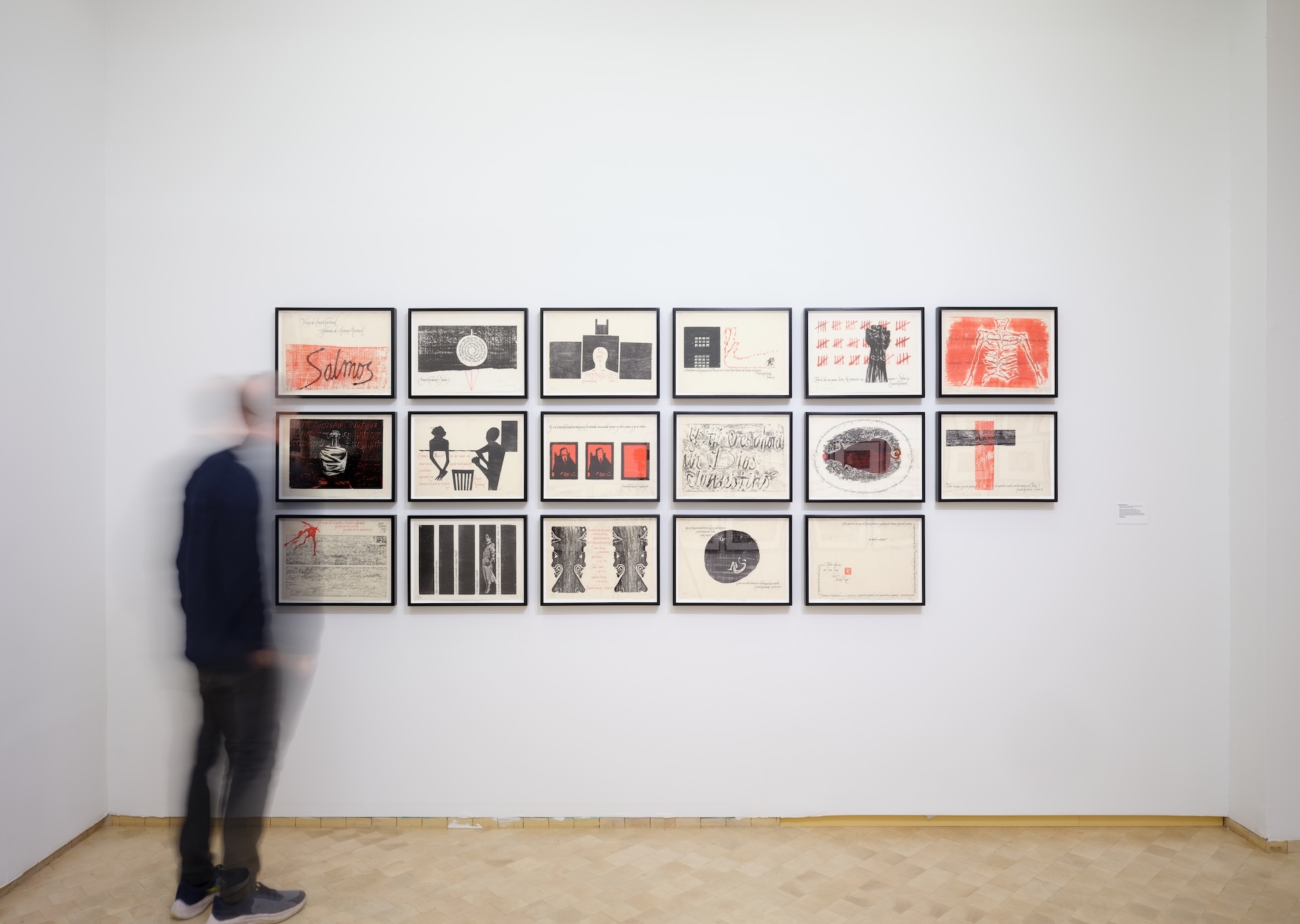 Person looking at three rows of framed works on a wall.