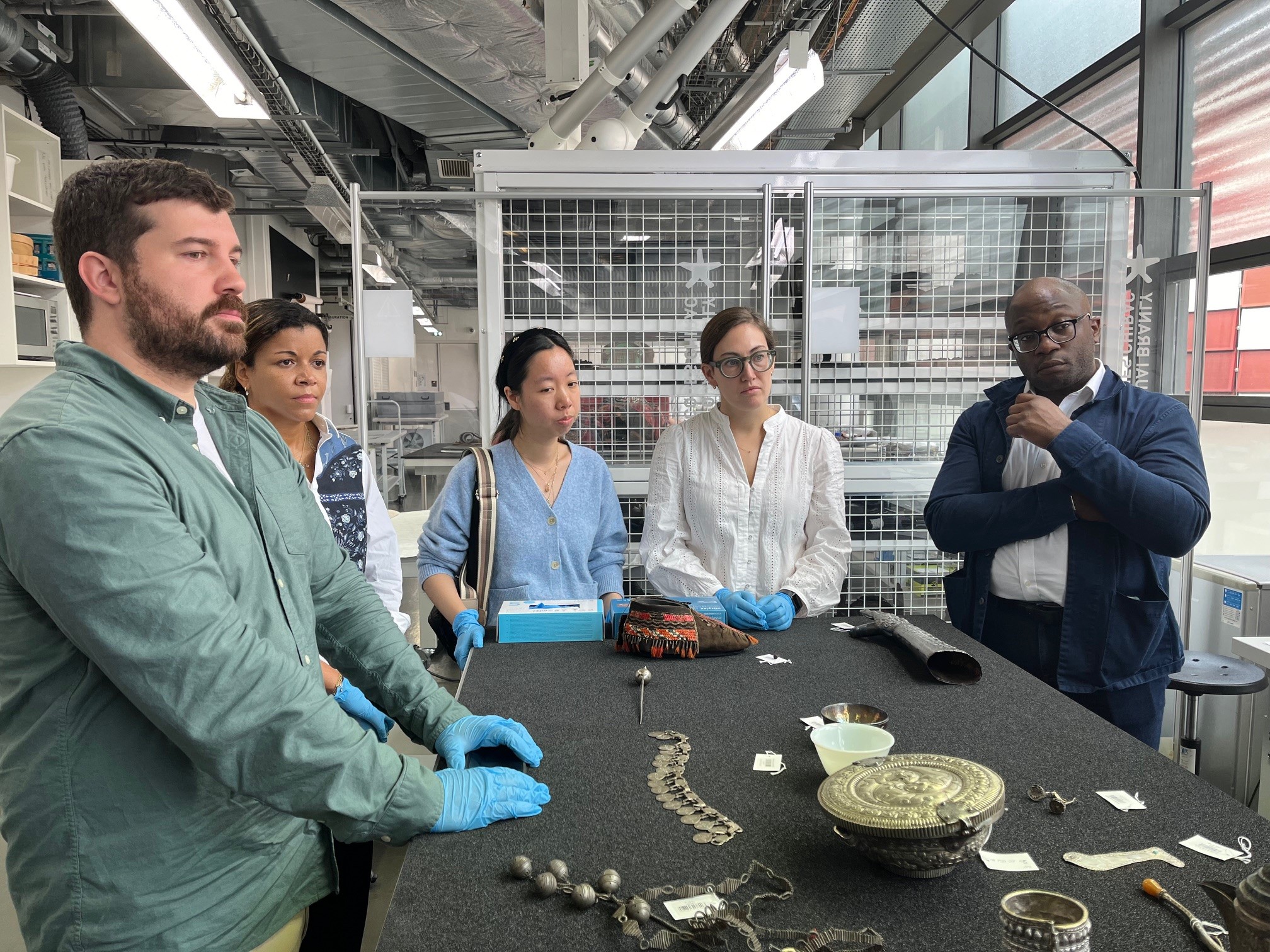 Five people standing around a table looking at objects made of silver.