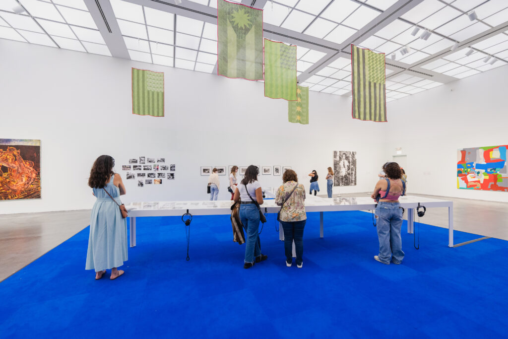 People standing in a large gallery space with blue carpet and white walls. Artwork is hanging on the walls and the center of the room has many vitrines.