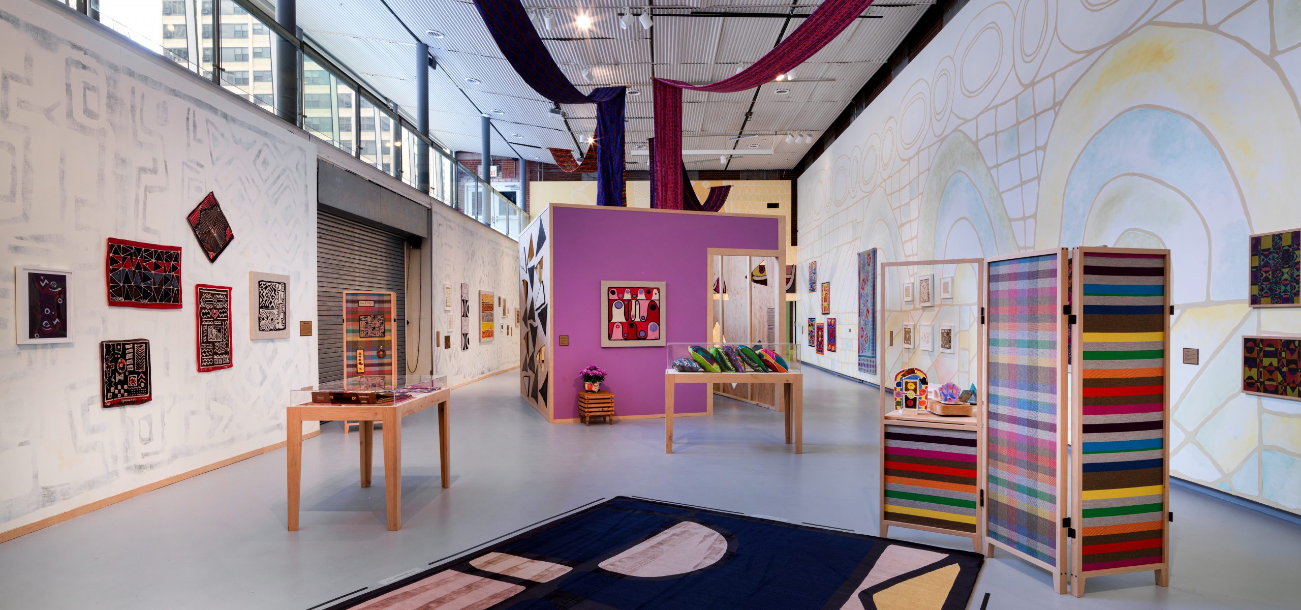 Installation of colorful textiles and objects in a long gallery space. The walls are each decorated with different painted patterns.
