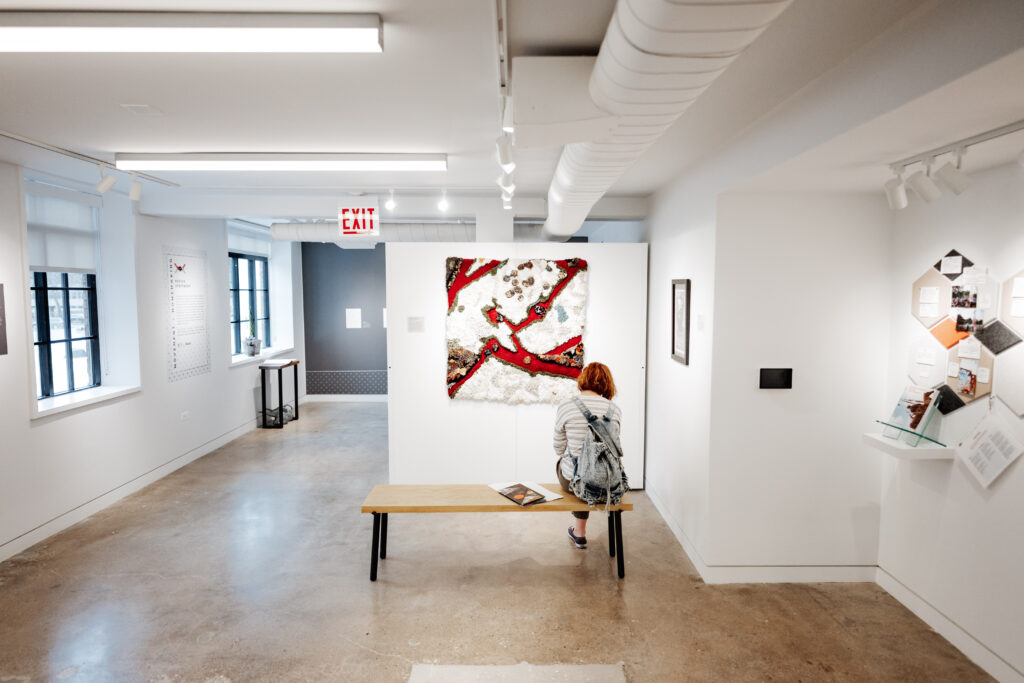 Woman seated on a bench in a gallery looking at a work of art in red and white installed on a gallery wall.