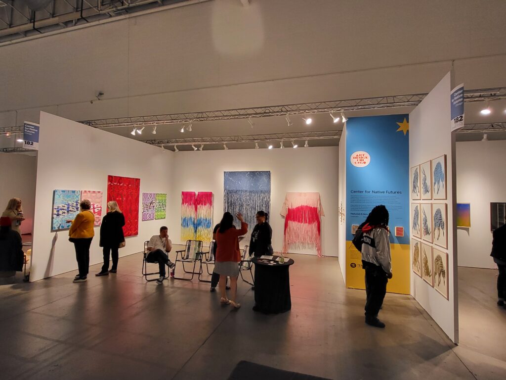 Center for Native Futures installation at an art exposition. Colorful objects are installed on white walls, with one wall with didactics painted blue and yellow.