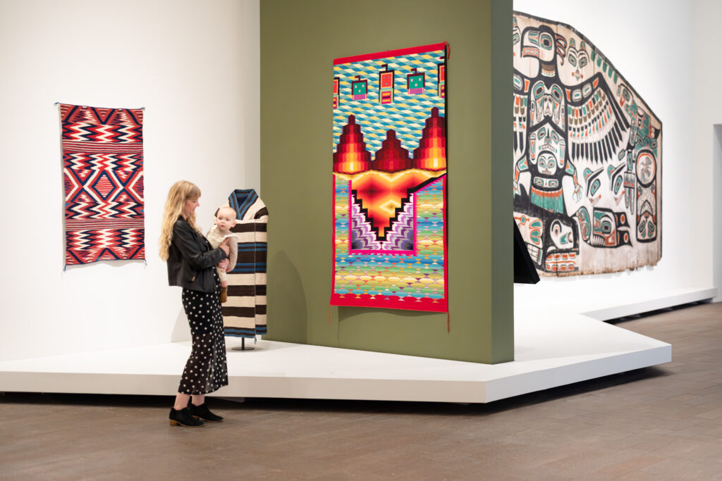 A woman holding a baby standing in front of textiles in a museum display.