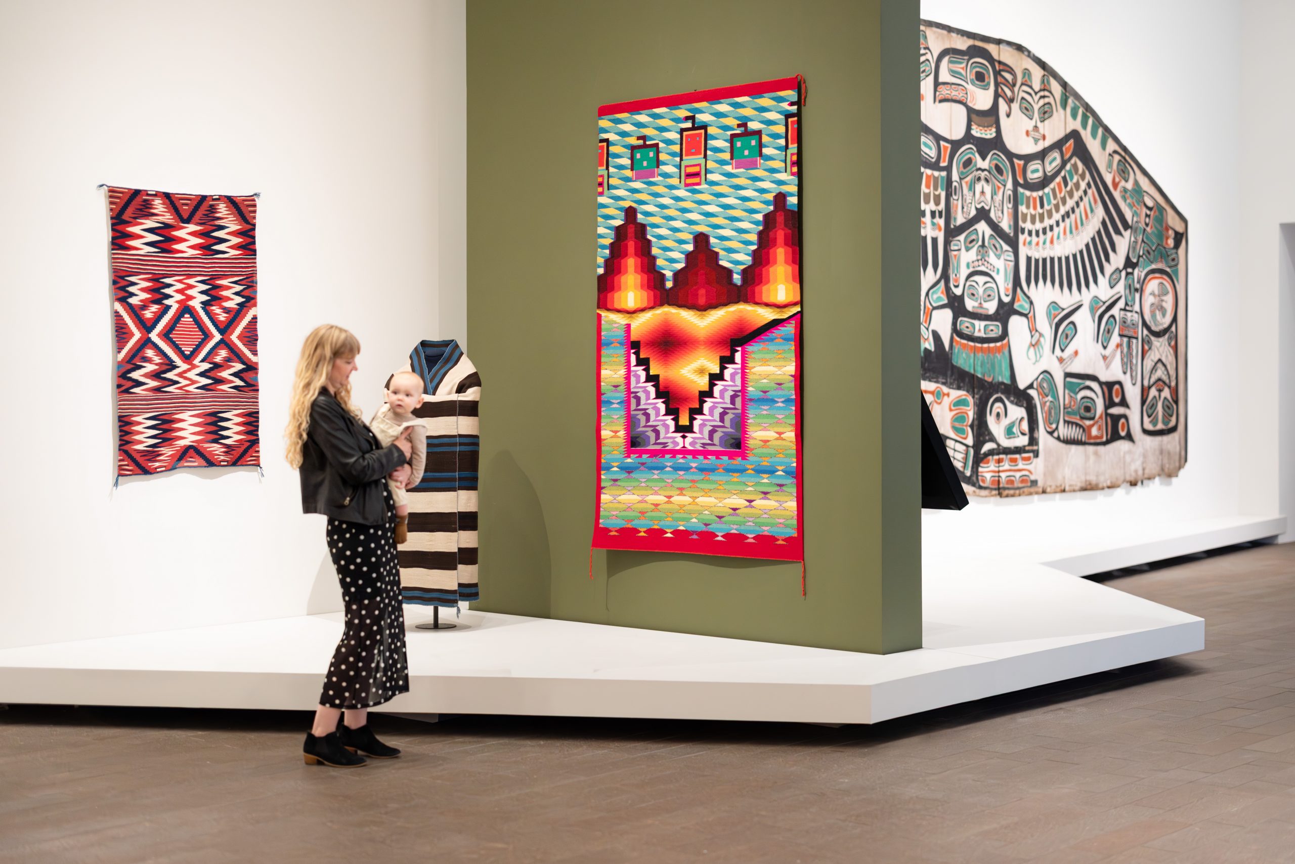 A woman holding a baby standing in front of textiles in a museum display.
