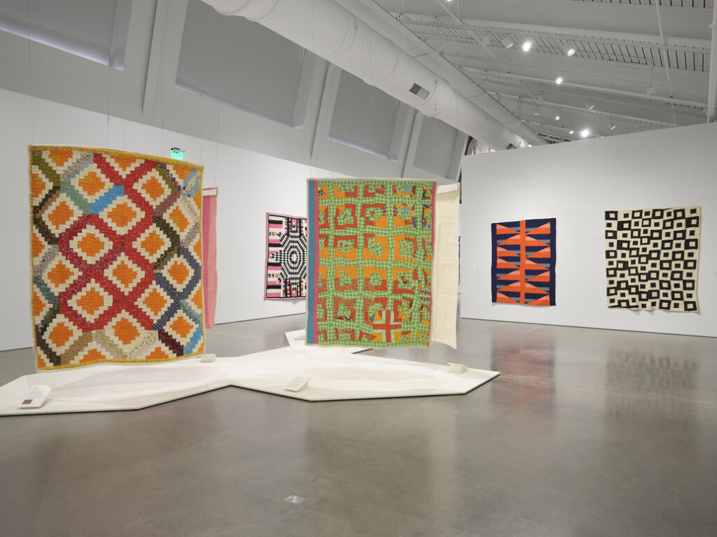 Installation view of colorful quilts suspended from the ceiling and installed on walls.